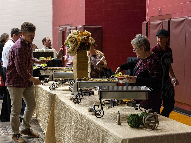 People serve themselves food from a buffet table at an indoor event. The table is covered with a beige cloth and decorated with flowers. Several trays of food are set out, and attendees are filling their plates.