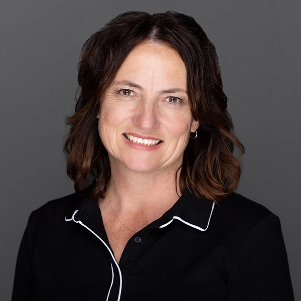 A woman with wavy brown hair, wearing a black collared shirt with white piping, smiles in front of a plain gray background.