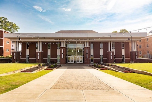 A brick academic building with large windows and columns at the entrance, surrounded by green lawns and sidewalks, under a blue sky. The sign reads Hope Barber Shull Academic Resource Center.