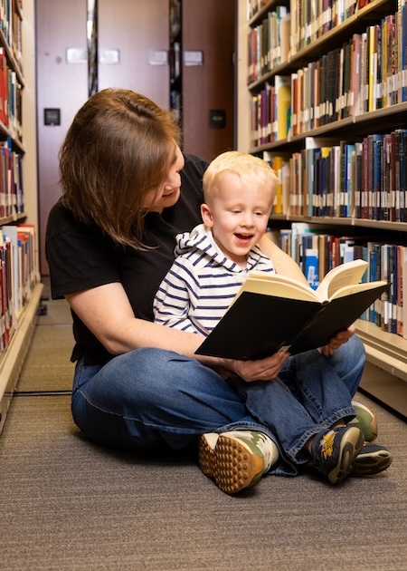 A woman and a young boy sit on the floor between library shelves, smiling as they read a book together. The woman holds the open book while the child looks at the pages with excitement.
