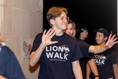 A group of young people wearing “Lion Walk” T-shirts smile and wave as they walk together at night. One person in the foreground raises a hand, appearing to greet or high-five someone.