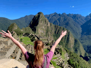 A person with long hair, arms raised, faces the ancient Inca ruins of Machu Picchu in Peru. Green mountains and a clear blue sky are visible in the background.