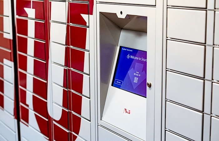 A close-up of a smart locker system featuring a touchscreen displaying a “Welcome to Smart” message and a QR code, with white locker doors and red text partially visible in the background.