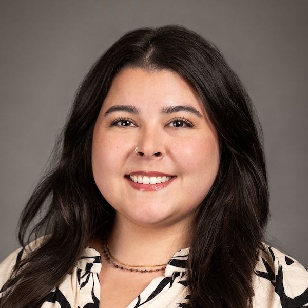 A woman with straight dark hair, wearing a brown and black floral-patterned blouse, smiles in front of a plain gray background.