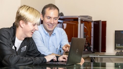 Two people sit at a table, smiling and looking at a laptop screen together. One is pointing at the screen, suggesting a collaborative or instructional setting. Theres technical equipment in the background.