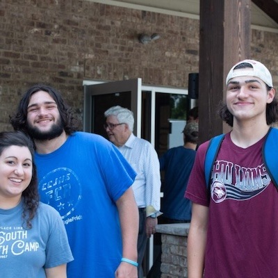 Three young people, two male and one female, are smiling outdoors in front of a brick building. The males have long hair and are wearing casual t-shirts; one has a backpack and baseball cap. An older man stands in the background.