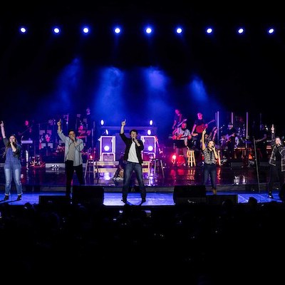 Five singers stand on a stage with one arm raised, performing in front of a live band under bright blue and purple stage lights. The audience is visible in silhouette at the bottom.