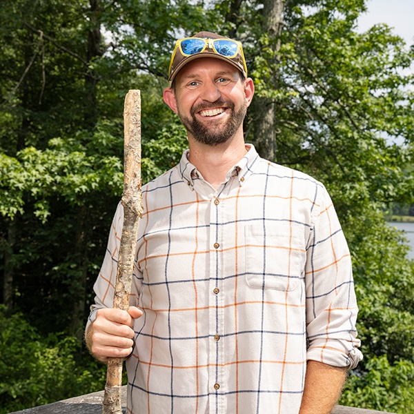 A smiling man in a plaid shirt and cap holds a walking stick while standing outdoors on a wooden deck, with green trees and a lake in the background.