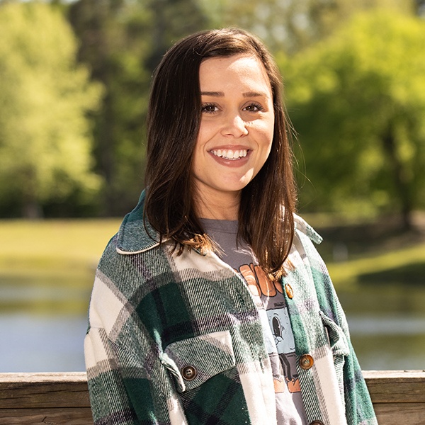 A young woman with straight brown hair smiles outdoors, wearing a green and white plaid jacket over a graphic t-shirt. Green trees and a pond are visible in the blurred background.