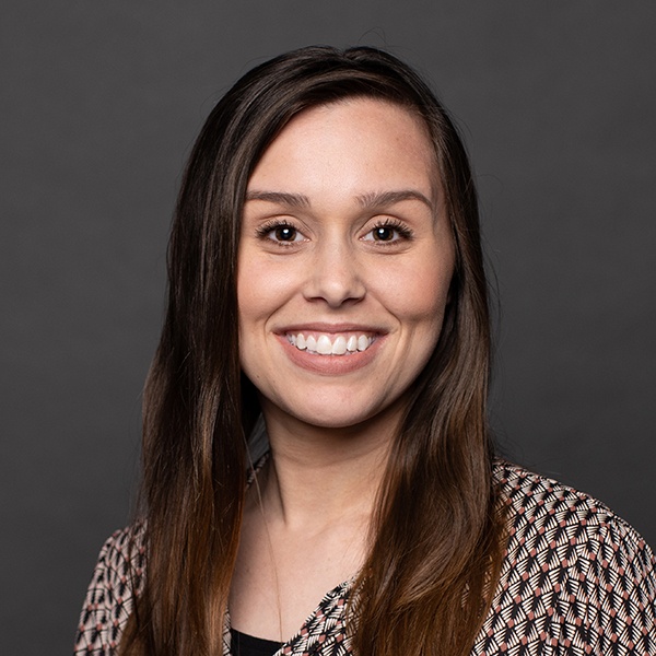 A young woman with long brown hair smiles at the camera. She is wearing a patterned blouse and is posed in front of a plain dark gray background.