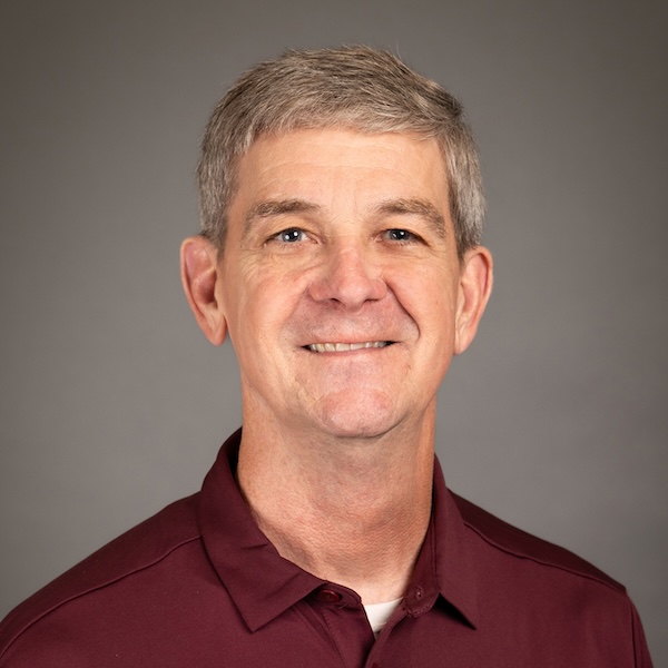 A middle-aged man with short gray hair smiles at the camera. He is wearing a gray zip-up sweater over a black shirt, and the background is plain and dark.