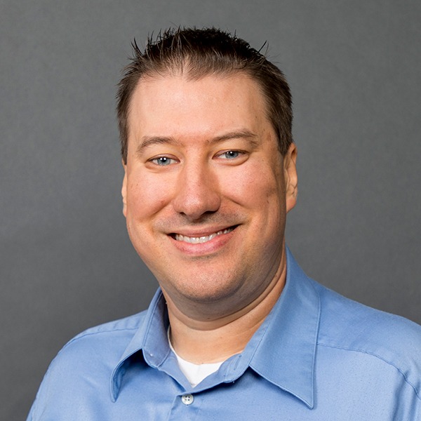 A smiling man with short brown hair wearing a light blue collared shirt, standing in front of a plain gray background.