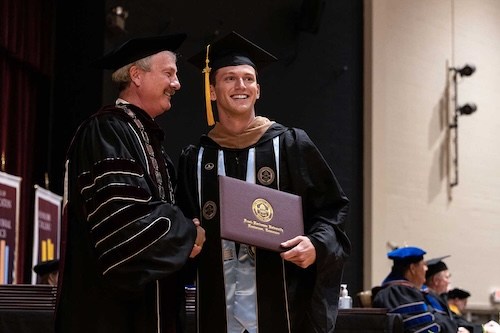 A graduate in a cap and gown smiles while shaking hands with a faculty member on stage and holding a diploma during a graduation ceremony. Other faculty members are seated in the background.