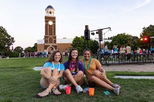 Three young women sit on grass smiling at the camera, with colorful cups in front of them. Behind them is a brick clock tower and a crowd gathered outdoors on a sunny day.