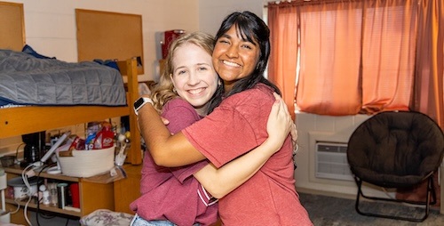 Two young women smiling and hugging each other in a cozy dorm room with bunk beds, a chair, and a window with pink curtains in the background.