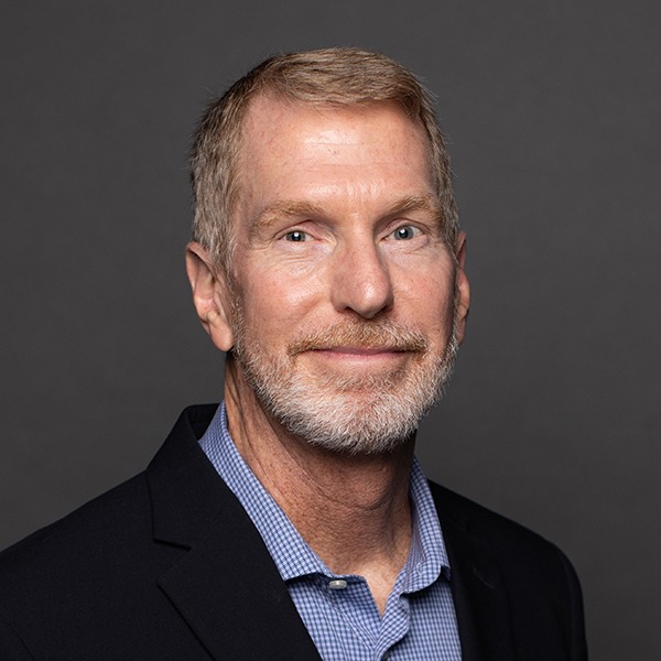A middle-aged man with short light hair and a trimmed beard, wearing a dark blazer over a blue checkered shirt, smiles slightly against a plain dark gray background.