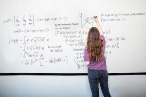 A person with long hair, wearing a purple shirt and jeans, writes complex mathematical equations and graphs on a large whiteboard. The board is filled with formulas, integrals, and diagrams.