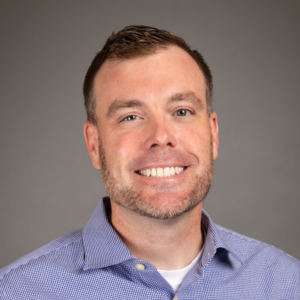 A smiling man with short hair and a trimmed beard, wearing a light-colored, checkered button-up shirt over a white undershirt, against a plain gray background.
