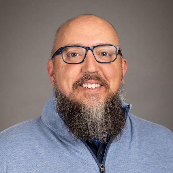 A man with a bald head, glasses, and a long beard is smiling. He is wearing a blue checkered suit jacket over a light blue collared shirt, with a plain gray background.