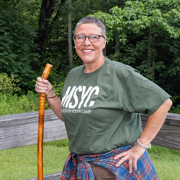 A smiling woman with short gray hair, glasses, and an MSYC t-shirt stands outside on a wooden deck holding a walking stick. She has a plaid shirt tied around her waist and green trees in the background.