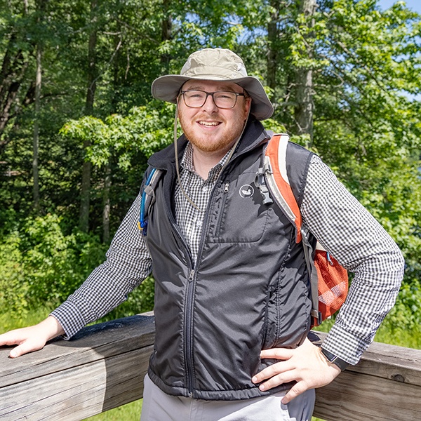A man wearing glasses, a sun hat, a black vest, and a checkered shirt stands outdoors by a wooden railing. He is smiling and has a backpack, with trees and greenery in the background.