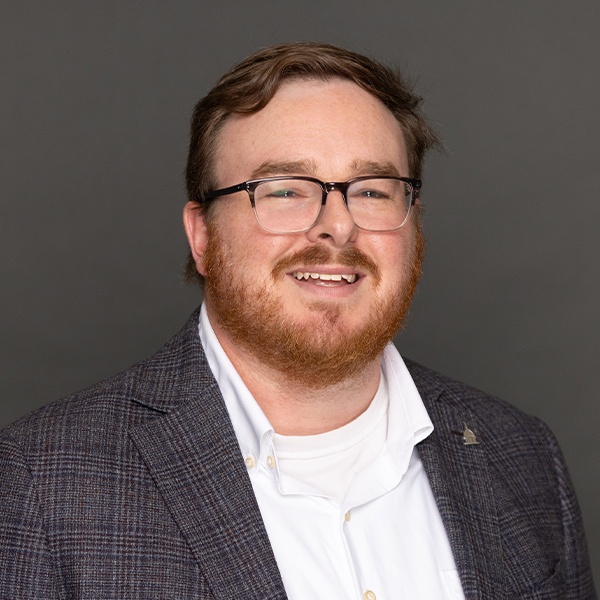 A man with short brown hair, a beard, and glasses is smiling. He is wearing a gray plaid blazer over a white collared shirt and a white undershirt. The background is plain and gray.