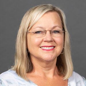 A woman with shoulder-length blonde hair, wearing glasses and a light patterned top, smiles at the camera against a plain gray background.