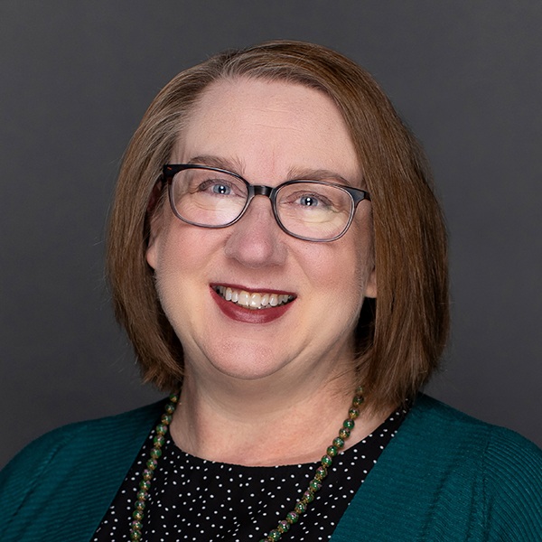A woman with shoulder-length light brown hair, glasses, and a dark green cardigan over a black polka dot top, smiling in front of a dark gray background. She is also wearing a beaded necklace.