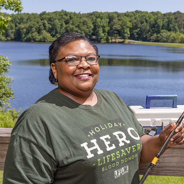 A smiling woman wearing glasses and a green Holiday Hero Lifesaver Blood Donor t-shirt stands holding a fishing rod in front of a scenic lake, with trees and a tackle box in the background.