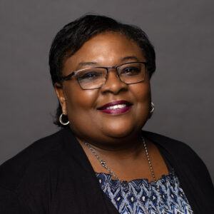 A woman with short dark hair, glasses, and hoop earrings smiles at the camera. She is wearing a patterned top, a black cardigan, a silver necklace, and lipstick, against a plain dark background.