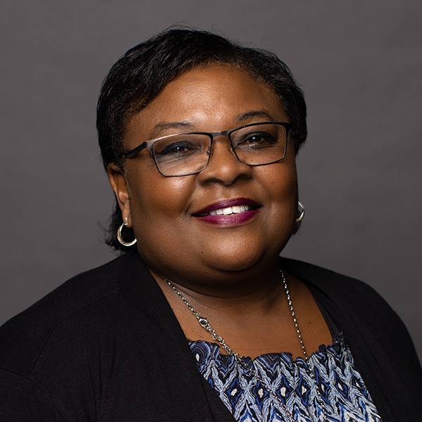 A woman with short dark hair, glasses, and hoop earrings smiles at the camera. She is wearing a patterned top, a black cardigan, a silver necklace, and lipstick, against a plain dark background.