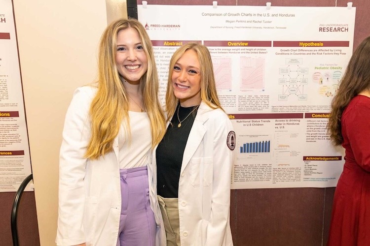 Two women in white lab coats smile in front of a scientific research poster about growth charts in the U.S. and Honduras. The poster contains sections like Abstract, Overview, and Hypothesis.