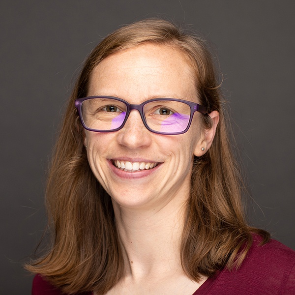 A smiling woman with straight, light brown hair, wearing purple glasses and a maroon top, poses in front of a plain dark background.
