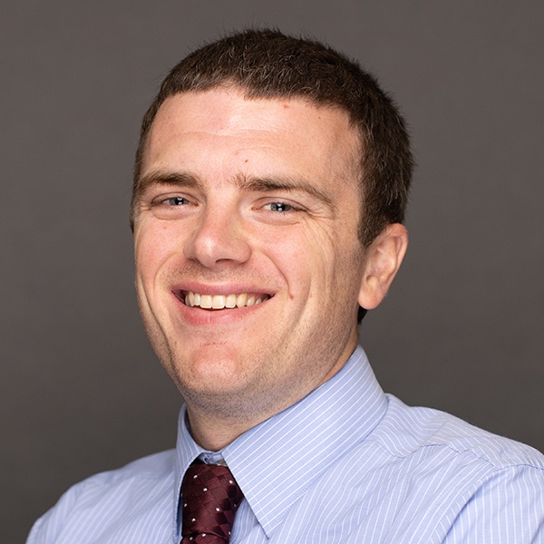 A man with short brown hair, wearing a light blue striped shirt and a maroon tie with small white dots, smiles in front of a plain dark grey background.