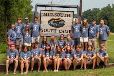 A group of young adults and teens wearing matching blue T-shirts pose and smile in front of a wooden Mid-South Youth Camp sign outdoors on a sunny day.