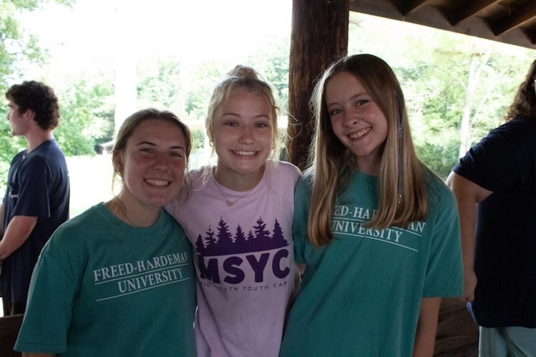 Three smiling teenage girls stand close together outdoors, two wearing teal Freed-Hardeman University shirts and one wearing a lavender MSYC shirt. Other people and trees are visible in the background.