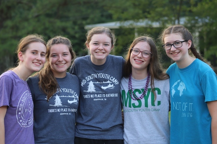 Five smiling teenage girls stand outdoors, close together, wearing casual T-shirts with Mid South Youth Camp and camp-themed designs. Trees and greenery are visible in the blurred background.