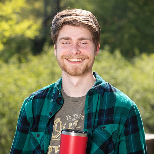 A young man with light brown hair and a beard smiles outdoors, wearing a green plaid shirt over a graphic tee, holding a red travel mug. Green foliage and trees are blurred in the background.
