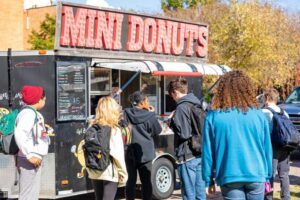 A group of students with backpacks stand in line at a black food truck with a large red MINI DONUTS sign on top. Trees with autumn leaves and a brick building are visible in the background.
