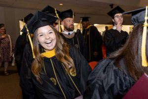 A young woman in a graduation cap and gown smiles and laughs while standing among other graduates indoors during a commencement ceremony.