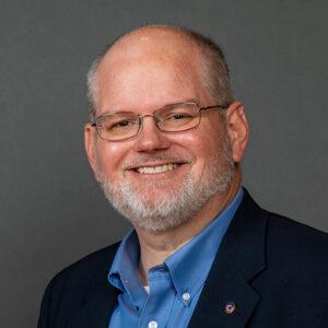 A middle-aged man with glasses, a gray beard, and a friendly smile wears a blue dress shirt and dark blazer, posing against a plain gray background.