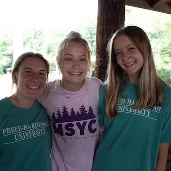 Three smiling teenage girls stand close together outdoors. Two wear matching teal Freed-Hardeman University shirts, and the one in the middle wears a purple MSYC camp shirt. Trees and sunlight are visible in the background.