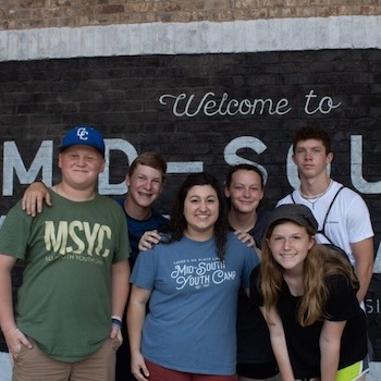 Six smiling teenagers stand in front of a black and white wall that says “Welcome to Mid-South.” They are posing closely together, some with arms around each other, and appear happy and casual.