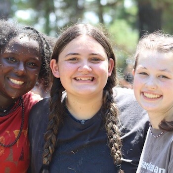 Three teenage girls stand close together outdoors, smiling brightly at the camera. The sunlight highlights their faces, and trees are visible in the blurred background. They appear happy and relaxed.