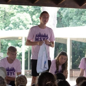 A young man in a pink MSYC shirt stands and speaks to a group outdoors, holding a towel. Two others in matching shirts sit nearby, with trees and a building in the background.