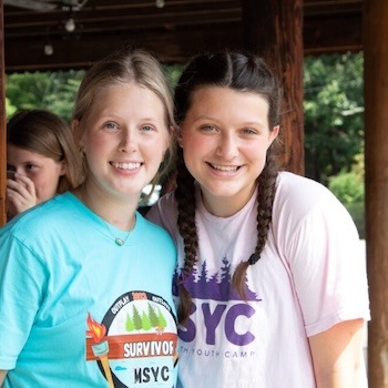 Two smiling teenage girls stand close together outdoors. One wears a blue SURVIVOR MSYC shirt, and the other wears a white MSYC Youth Camp shirt with purple graphics. Another girl stands in the background, partially visible.