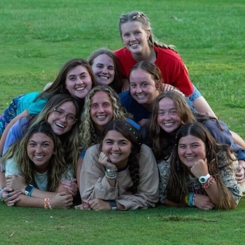 A group of ten young women are smiling and lying in a pyramid formation on green grass outdoors, looking happy and relaxed.