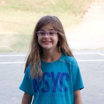 A young girl with long brown hair and glasses smiles while standing outdoors on a paved area, wearing a blue MSYC T-shirt. Grass and a path are visible in the background.