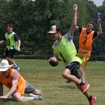 Four young men play a game of flag football outside. One player in a green vest runs with the ball while others, wearing orange vests, chase him. Another player in white has fallen on the grass. Trees are visible in the background.