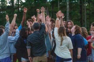 A large group of people stands in a circle outdoors with their hands raised together in the center, surrounded by trees, showing unity and enthusiasm.